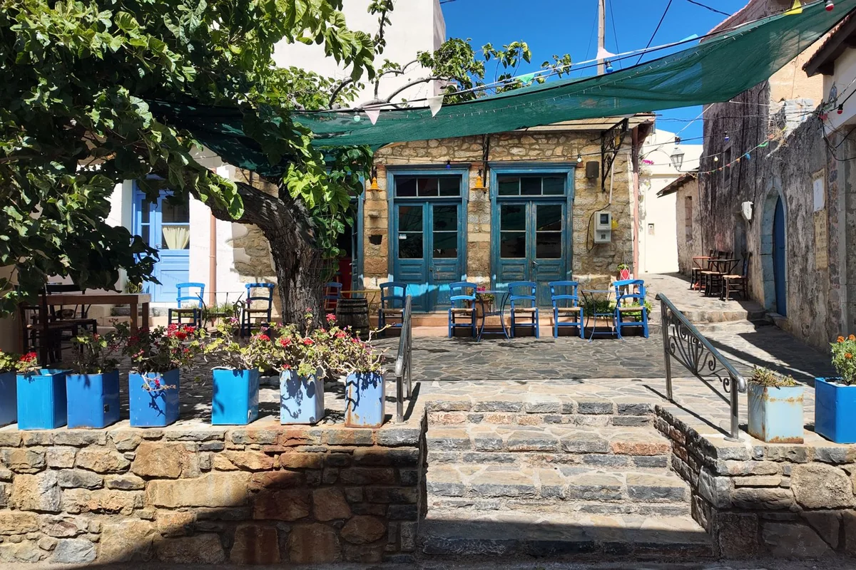 Kamenná taverna s natřenými modrými židlemi, stínicí markýzou a světelnými řetězy — typická tradiční řecká taverna, nikoli skutečné místo konání